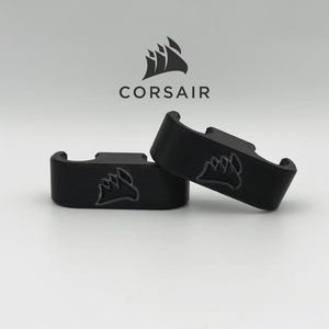 May include: Two black, curved brackets with the Corsair logo, a stylized ship, are displayed against a white background. The word "CORSAIR" is above the brackets. The brackets are likely computer accessories.