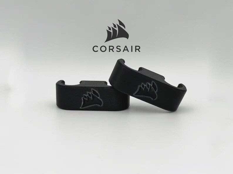 May include: Two black, curved brackets with the Corsair logo, a stylized ship, are displayed against a white background. The word "CORSAIR" is above the brackets. The brackets are likely computer accessories.