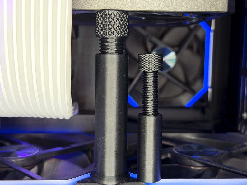Puede incluir: Primer plano de dos tornillos negros roscados con tapas texturizadas, probablemente para hardware de computadora. Los tornillos son de diferentes tama&ntilde;os y est&aacute;n colocados frente a un ventilador de computadora y otros componentes internos. Iluminaci&oacute;n LED azul visible.