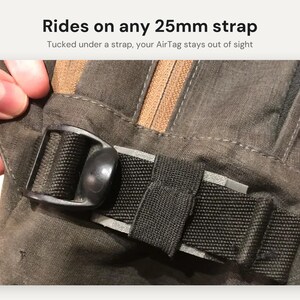 May include: A black plastic AirTag holder attached to a dark gray fabric strap. The text reads: Rides on any 25mm strap. Tucked under a strap, your AirTag stays out of sight. The holder features a black buckle mechanism.