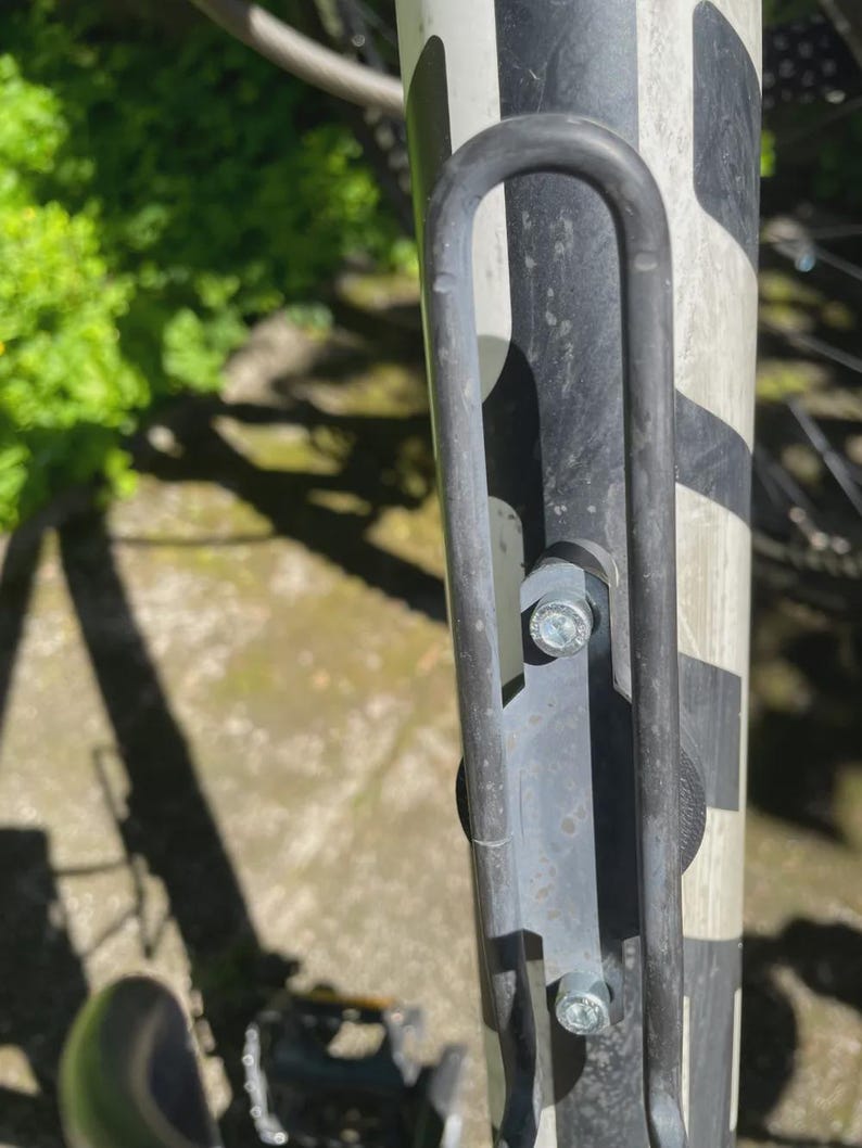 May include: Close-up of a bicycle frame with a black and white pattern. A grey metal water bottle cage is attached with silver bolts. The background shows green foliage and sunlight.