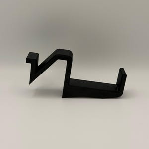 Op de afbeelding: Een zwart, hoekig object, mogelijk een haak of houder, met een unieke vorm. Het ontwerp heeft een zigzag-element verbonden met een horizontale basis met een verhoogde rand. Het object is gemaakt van een solide, ondoorzichtig materiaal en ligt op een witte ondergrond.