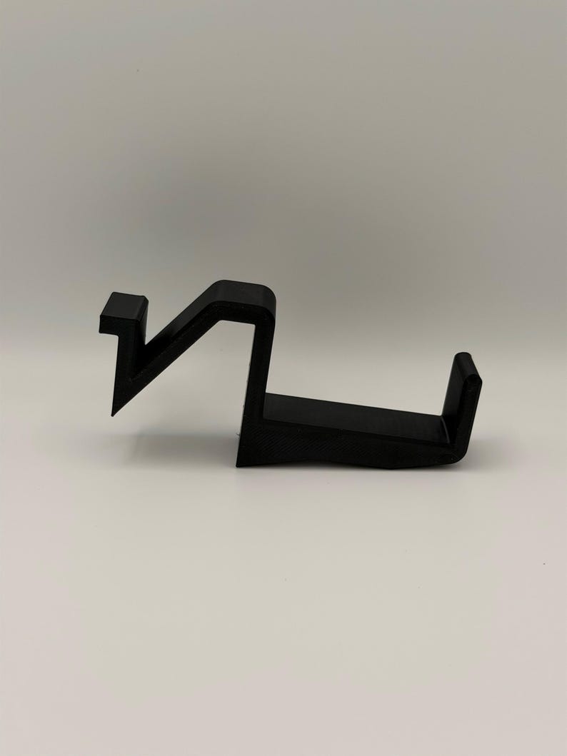 Op de afbeelding: Een zwart, hoekig object, mogelijk een haak of houder, met een unieke vorm. Het ontwerp heeft een zigzag-element verbonden met een horizontale basis met een verhoogde rand. Het object is gemaakt van een solide, ondoorzichtig materiaal en ligt op een witte ondergrond.