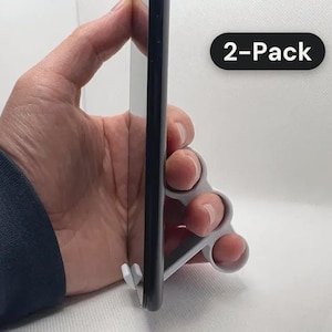 May include: A white plastic smartphone grip featuring three circular finger holes, attached to the side of a black smartphone. The accessory is labeled with the text 2-Pack, indicating a set of two units for mobile device handling.