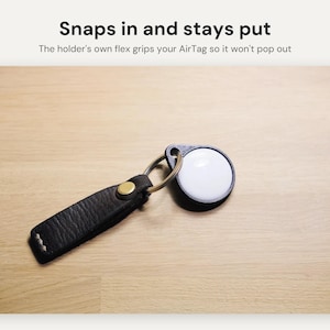 May include: A black textured leather keychain holder with a brass snap button and a metal ring, securing a white circular AirTag. The text above reads: Snaps in and stays put. The holder's own flex grips your AirTag so it won't pop out.