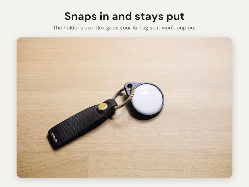 May include: A black textured leather keychain holder with a brass snap button and a metal ring, securing a white circular AirTag. The text above reads: Snaps in and stays put. The holder's own flex grips your AirTag so it won't pop out.