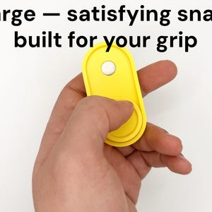 May include: A bright yellow, oval-shaped grip accessory with a white circular button. The text "Large - satisfying snap, built for your grip" is displayed in black. The accessory is held in a hand against a white background.