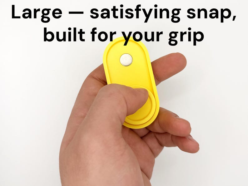 May include: A bright yellow, oval-shaped grip accessory with a white circular button. The text "Large - satisfying snap, built for your grip" is displayed in black. The accessory is held in a hand against a white background.