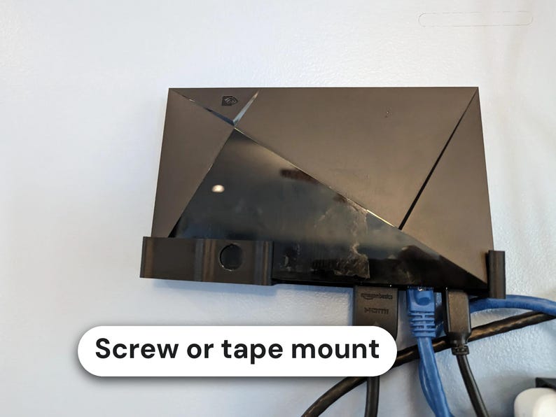 May include: A black plastic wall mount holding a black media streaming device. The mount features a rectangular base with side supports and a circular opening. The text "Screw or tape mount" is visible on a white label below the device.