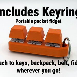 K&ouml;nnte beinhalten: Orangenes, tragbares Taschen-Fidget mit drei quadratischen Tasten und einem Schl&uuml;sselring. Der Text auf dem Bild lautet "Includes Keyring" und "Attach to keys, backpack, belt, fidget wherever you go!"