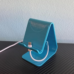 May include: A teal-colored phone stand with a curved design. A white charging cable with a pink connector is inserted into the stand. The stand is made of plastic and is designed to hold a phone upright.