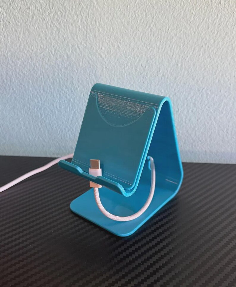 May include: A teal-colored phone stand with a curved design. A white charging cable with a pink connector is inserted into the stand. The stand is made of plastic and is designed to hold a phone upright.