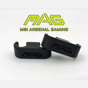 May include: Two black MAG gaming accessories are displayed against a white background. The accessories have the word "MAG" embossed on them. The logo "MAG MSI ARSENAL GAMING" is above the accessories in yellow and black.