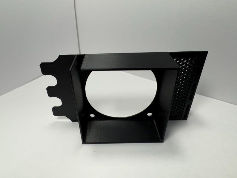 May include: A black, angular bracket with a circular opening and a perforated side panel. The bracket has a series of cutouts on one end and two small holes near the base of the circular opening. The object is set against a white background.