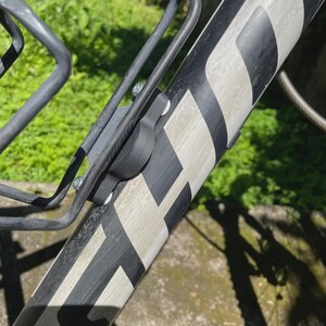 May include: A black and grey bicycle frame with a water bottle cage attached. The frame features a white and black pattern. The water bottle cage is metal and secured with screws. The background shows blurred green foliage.