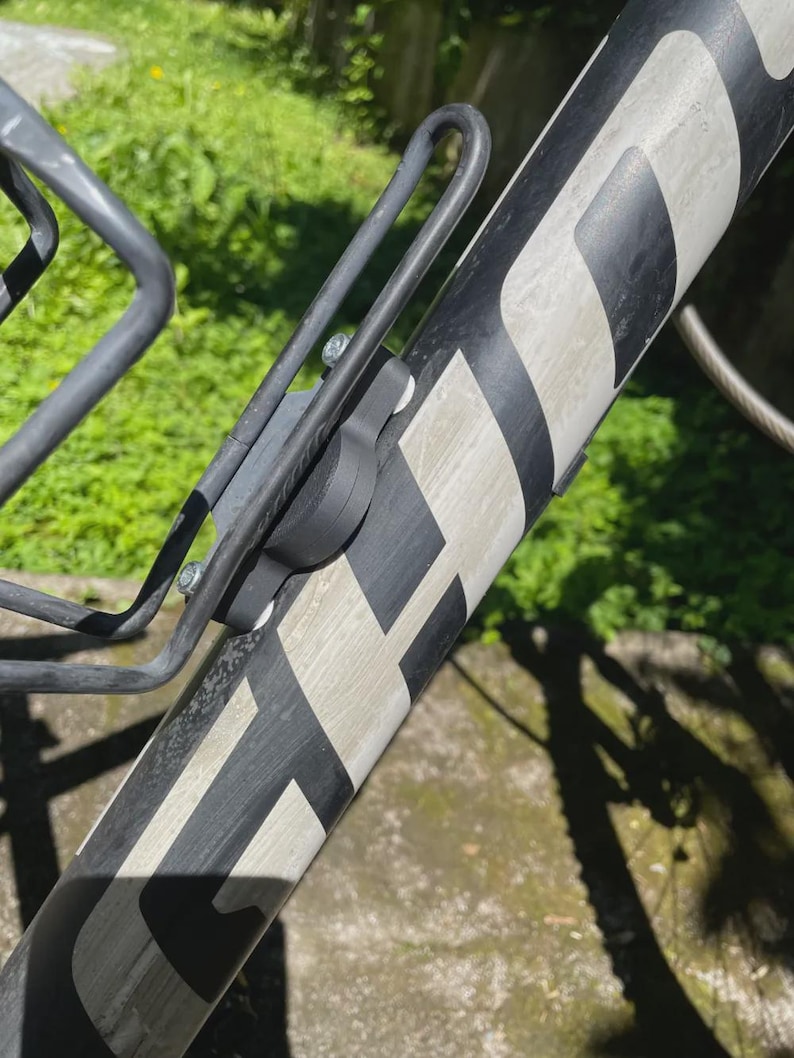 May include: A black and grey bicycle frame with a water bottle cage attached. The frame features a white and black pattern. The water bottle cage is metal and secured with screws. The background shows blurred green foliage.