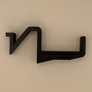 Op de afbeelding: Een zwarte, geometrische wandplank met een uniek, hoekig ontwerp. De plank is gemaakt van een textuurrijk materiaal en heeft een reeks verbonden, rechte lijnen en rechte hoeken. De plank is ontworpen om items vast te houden en is bevestigd aan een neutraal gekleurde muur.