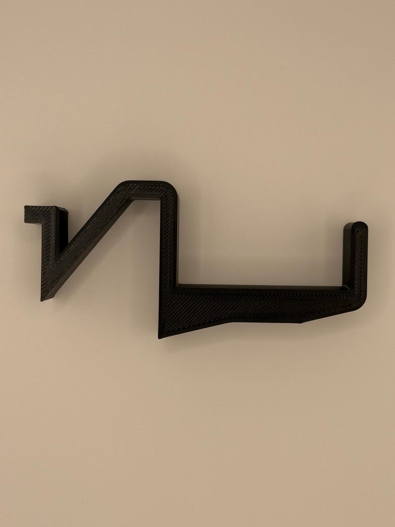 Op de afbeelding: Een zwarte, geometrische wandplank met een uniek, hoekig ontwerp. De plank is gemaakt van een textuurrijk materiaal en heeft een reeks verbonden, rechte lijnen en rechte hoeken. De plank is ontworpen om items vast te houden en is bevestigd aan een neutraal gekleurde muur.