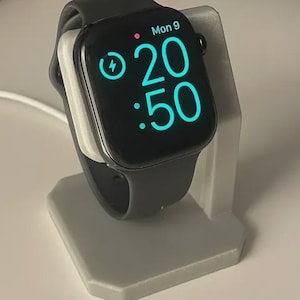 May include: A black smartwatch with a black strap rests on a grey charging stand. The watch face displays the time as 20:50 and the day as Mon 9. A lightning bolt icon indicates the watch is charging.