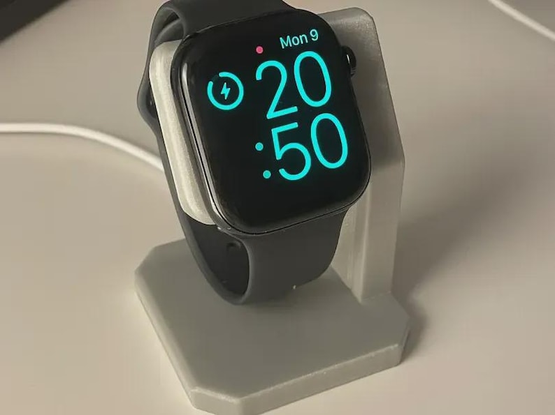 May include: A black smartwatch with a black strap rests on a grey charging stand. The watch face displays the time as 20:50 and the day as Mon 9. A lightning bolt icon indicates the watch is charging.