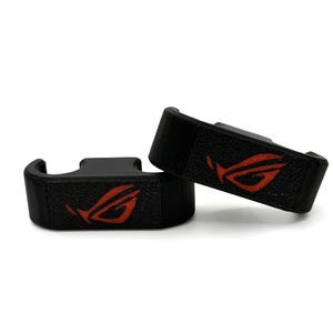 May include: Two black, textured gaming accessories with a red logo. The accessories are rectangular with a curved hook shape. The red logo is a stylized design. The items are set against a white background.