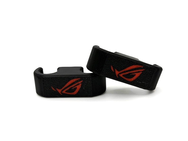 May include: Two black, textured gaming accessories with a red logo. The accessories are rectangular with a curved hook shape. The red logo is a stylized design. The items are set against a white background.