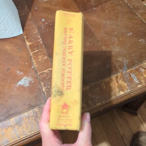 May include: A yellow hardcover book spine titled Harry Potter and the Deathly Hallows by J.K. Rowling. The spine features red text, a small red icon near the top, a red symbol near the bottom, and the Scholastic logo at the base of the spine.
