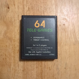 May include: A black Tele-Games 64 video game cartridge. The label features the text "64" in orange, "TELE-GAMES" in green, and game titles "HYPERSPACE" and "THRUST CONTROL". The cartridge is for 1 or 2 players and is compatible with joystick controllers.