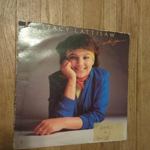 May include: Album cover for Stacy Lattisaw's "With You" album. The cover features a smiling woman in a blue jacket, yellow headband, and red scarf. The album cover is on a wooden surface.