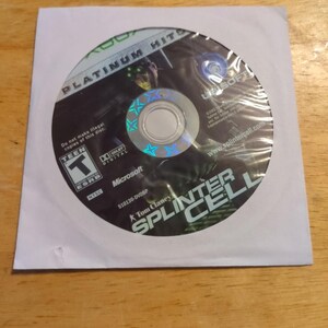 May include: A disc for the Xbox game "Tom Clancy's Splinter Cell" in a white paper sleeve. The disc is labeled "Platinum Hits" and features a character with green night vision goggles. The game is rated "Teen" by the ESRB.