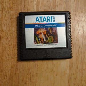May include: Black Atari 5200 Missile Command video game cartridge. The label features the Atari logo, game title, and a colorful graphic of a missile launch. The cartridge is rectangular with a ridged edge and is set against a wooden background.
