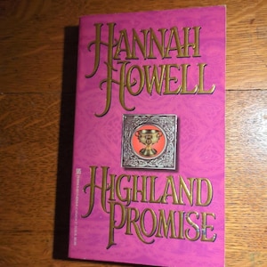 May include: A paperback book titled "Highland Promise" by Hannah Howell. The cover is a vibrant pink with gold lettering and a small illustration of a chalice. The book is resting on a wooden surface.