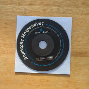 May include: A black CD with Greek text, including the name "Δημήτρης Μητροπάνος" and "BHMA RADIO" logo, is placed on a white paper square. The CD is centered on a light brown wooden surface.
