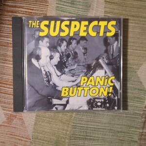 May include: A CD case featuring the album "Panic Button!" by "The Suspects." The cover art is a black and white photo of several men in a studio setting, with the album title in yellow text.