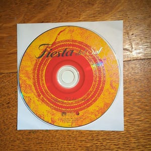 May include: A CD with a yellow and red design, featuring the text "Fiesta del Sol" in a decorative font. The CD is in a white paper sleeve, set against a wooden background.