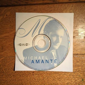 May include: A CD with a light blue image of a man and the name "MICHAEL AMANTE" printed in blue. The CD is on a white paper sleeve with the "MEDALIST" logo. The background is a wooden surface.