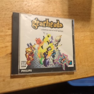 May include: A CD-ROM case for the game "Gearheads" featuring a colorful illustration of wind-up toys. The case has the Philips logo and the text "a furious war of wind-up toys". The case is on a wooden surface.