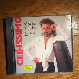 May include: CD case featuring Mischa Maisky and Daria Hovora. The cover has a portrait of a man in a white shirt and black pants, leaning against a dark case. The title "CELLISSIMO" is in red letters.