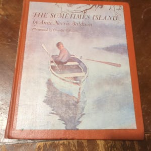 May include: A vintage book titled "The Sometimes Island" by Anne Norris Baldwin, illustrated by Charles Robinson. The cover features a watercolor illustration of a person rowing a boat on a lake. The book has a red-orange cover.