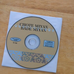May include: A compact disc with the text "CHOTE MIYAN BADE MIYAN" in black lettering. The disc is tan with blue and white decorative elements and a "Compact Disc Digital Audio" logo. It rests in a white paper sleeve.