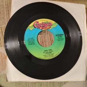 May include: A black vinyl record with a colorful label featuring the word "Chelsea" in pink and green. The record is titled "LOVE FIRE" by JIGSAW. The record is in a white paper sleeve.