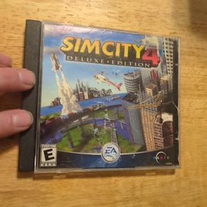 May include: SimCity 4 Deluxe Edition PC game in a clear plastic case. The cover art depicts a city with a rocket launch, a helicopter, and skyscrapers. The game is rated "E" for Everyone.