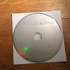 May include: A silver DVD disc with the title "The Curious Case Of Benjamin Button" printed in gold. The disc is in a white paper sleeve, set against a wooden background. The DVD logo and other details are visible.