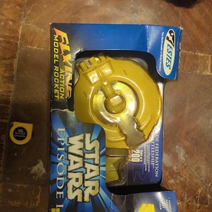 May include: A yellow and gray Star Wars Episode I model rocket in its original blue box. The box features the text "FLYING ACTION MODEL ROCKET" and "TRADE FEDERATION BATTLESHIP". The rocket is a toy.