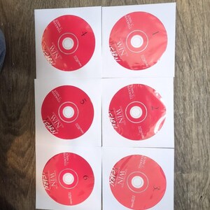 May include: Six red CDs with white paper sleeves are arranged on a wooden surface. Each CD has the text "WIN" and "LEARN" with the name "JOHN C MAXWELL". The CDs are numbered 1 through 6.