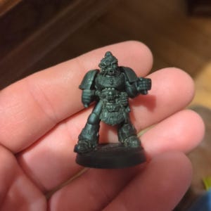 May include: A miniature, gray-colored figurine of a warrior in armor, standing on a circular base. The figure has detailed armor, a helmet, and a weapon. The figurine is likely for tabletop gaming or collecting.