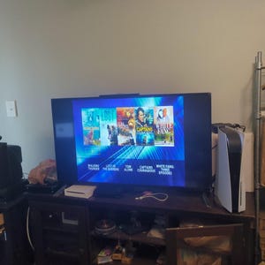 May include: A large flat-screen TV displays a menu with movie titles like "Lost" and "White Fang." A retro TV and a gaming console are on a dark wood entertainment center. A white and black gaming console is on the right.