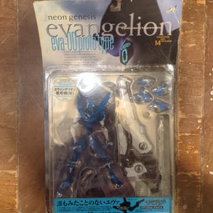 May include: Sealed action figure of the Eva-00 Proto Type from Neon Genesis Evangelion. The figure is blue and white, with multiple optional parts. The packaging includes text in English and Japanese.