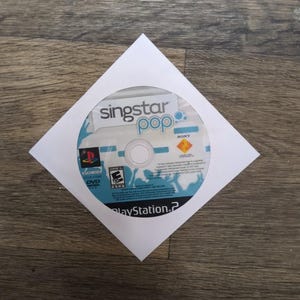 May include: A Singstar Pop PlayStation 2 DVD in a white paper sleeve. The disc has a blue and white design with the game title and PlayStation 2 logo. The disc is in a white paper sleeve on a wooden surface.