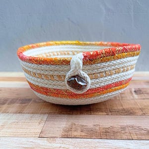 May include: A round bowl constructed from coiled rope featuring alternating bands of off-white, orange, and gold-flecked fabric. A single off-white rope loop is attached to the side, secured by a dark brown, polished, irregular-shaped stone bead.